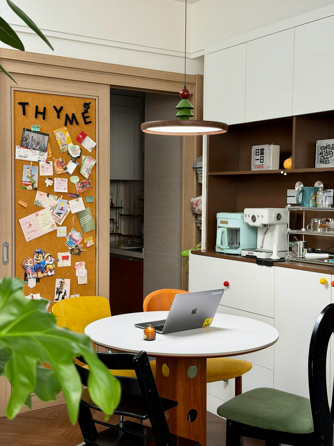 A laptop open on a home kitchen table beside a cork board of family photos